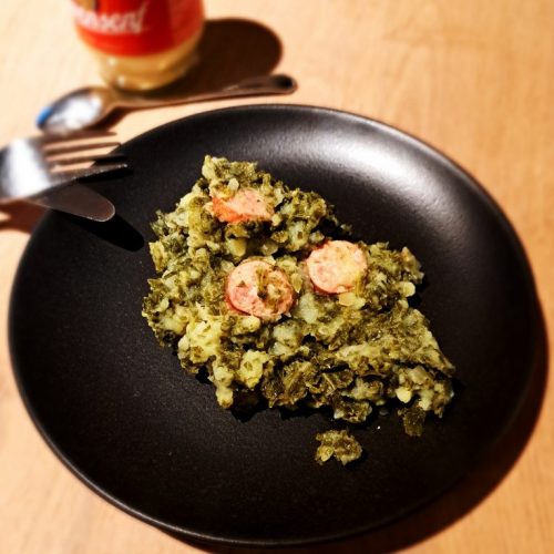 Grünkohl mit Kartoffeln und Mettwürste auf einem dunklen Teller. Besteck und ein Glas Senf im Hintergrund.